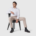 Man wearing beige slim-fit Jetsetter Tech Pant, sitting on a chair with a white shirt and black shoes.