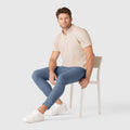 Man wearing Jetsetter Tech Pant Slim Fit in blue, seated on a chair, paired with a beige shirt and white sneakers; ideal for travel with wrinkle-free and stain-repellent features.